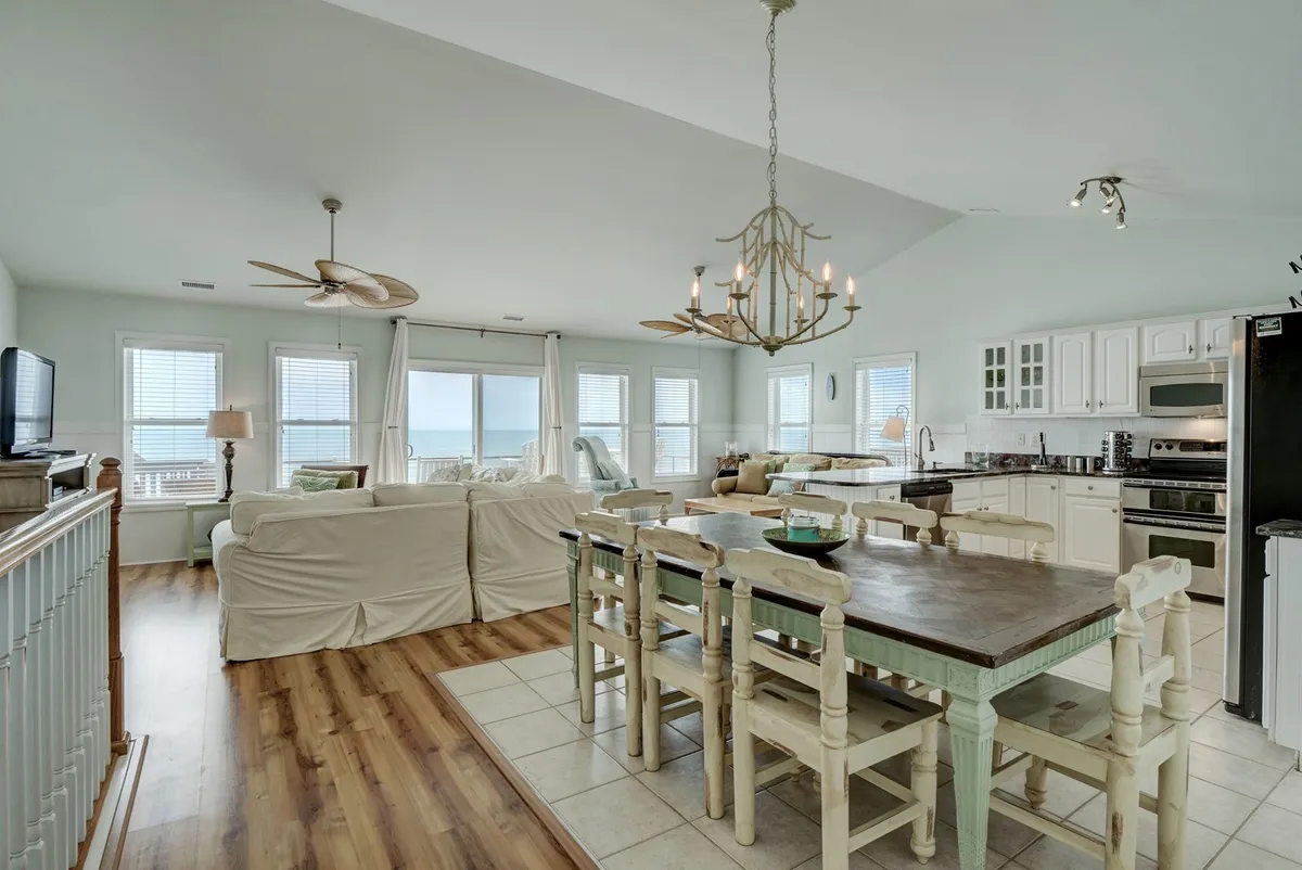 The dining area features a large dining table positioned near wide windows that offer glimpses of the ocean, with a visible kitchen area in the background. Decorative ceiling fans and natural light enhance the warm, inviting atmosphere, perfect for family meals and entertaining guests.