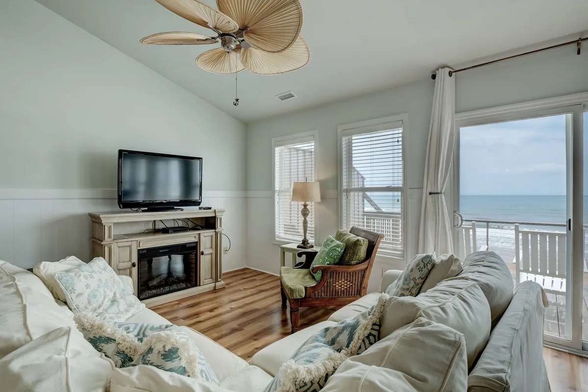 This image showcases a comfortable living room featuring a plush sofa with decorative pillows, large windows that frame a spectacular ocean view, a ceiling fan, and a television unit. The scene exudes a casual yet stylish atmosphere, with natural light flooding the space and highlighting the room’s airy feel.