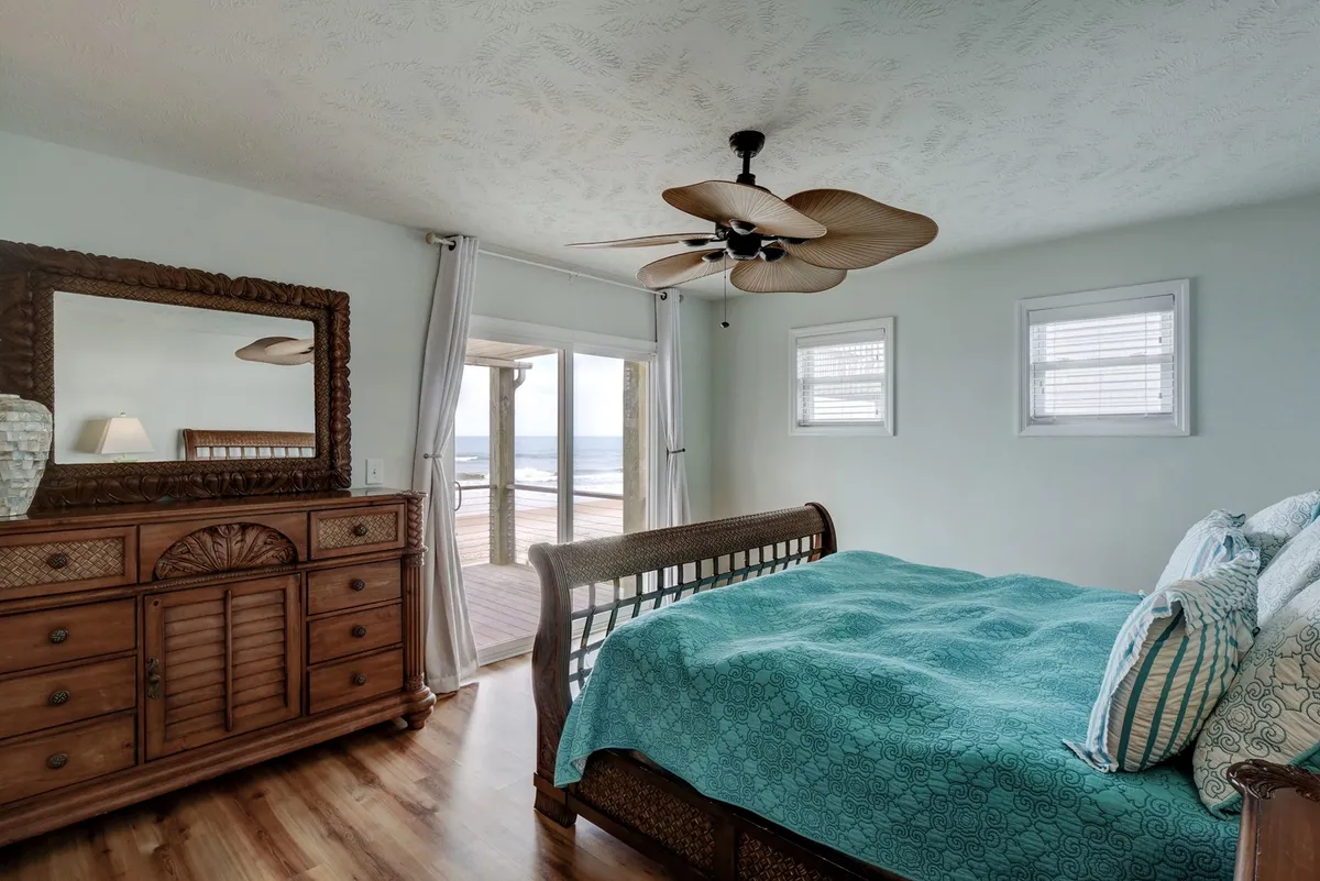A tranquil master bedroom with aqua bedding, a wooden dresser, large windows offering sweeping ocean views, and a ceiling fan overhead. The room is bathed in natural light and exudes a sense of calm and relaxation, perfectly positioned to take in the sounds and sights of the ocean from the comfort of bed.