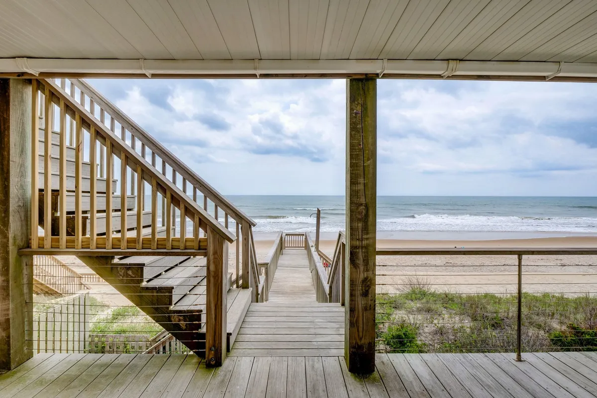 A wooden stairway leads down from the property to the sandy pathway and beach, with a panoramic view of the ocean inviting exploration. The scene suggests easy transitions between home and shore for all ages.