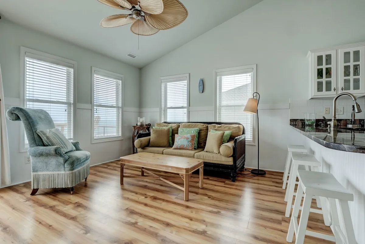 A stylish living area featuring a comfortable sofa and armchair set beside a dining area, with large windows that let in abundant light and offer glimpses of the natural surroundings. The decorative lamp and soft furnishings create a welcoming, intimate vibe, while the visible sunset light suggests the possibility of enjoying views of the horizon.