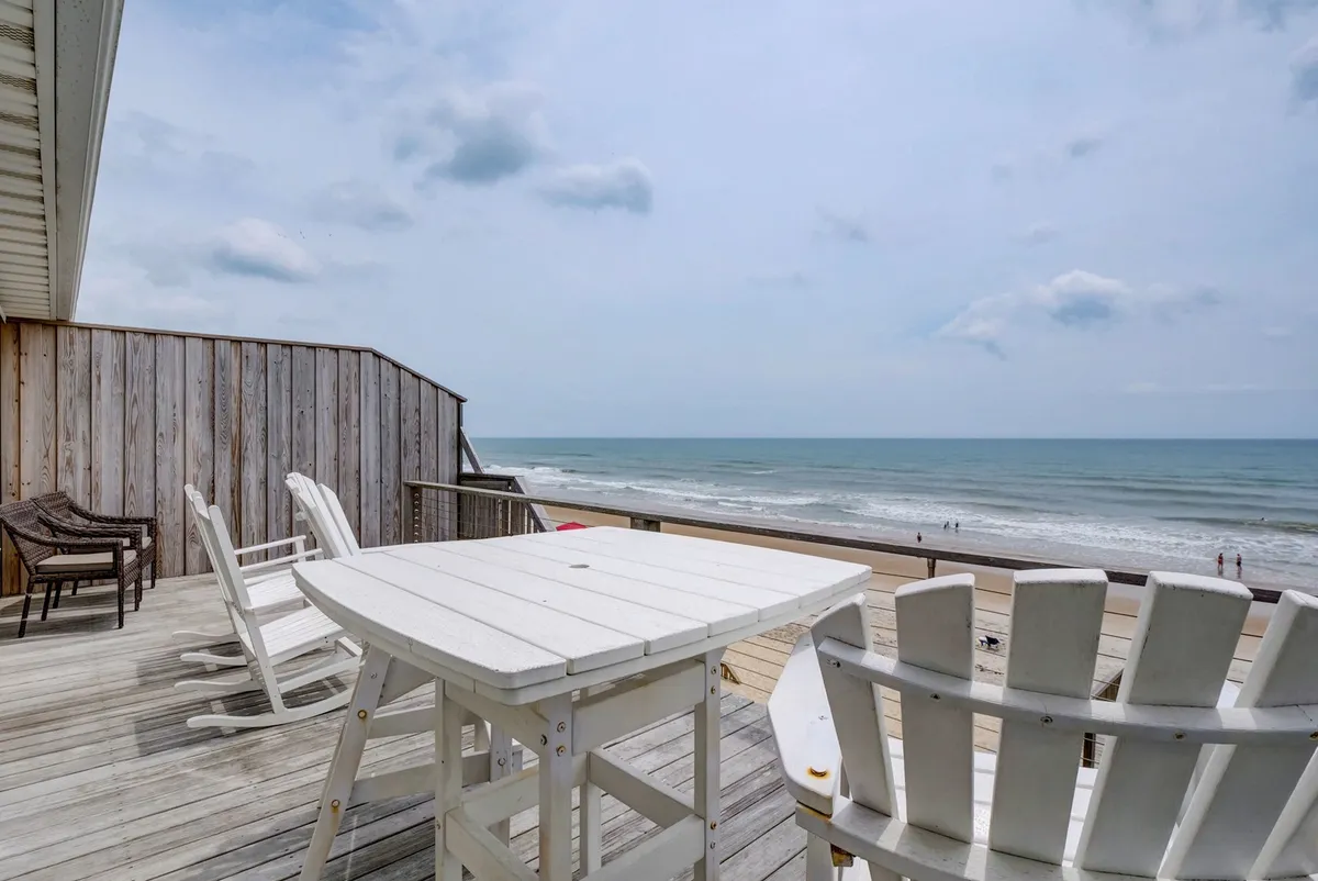 A serene oceanfront deck featuring comfortable deck chairs and a wooden table, all oriented toward the expansive ocean view. The space is ideal for sunbathing, enjoying meals, or gathering for cocktails, with the modern railing in view and the calming sound of waves implied.