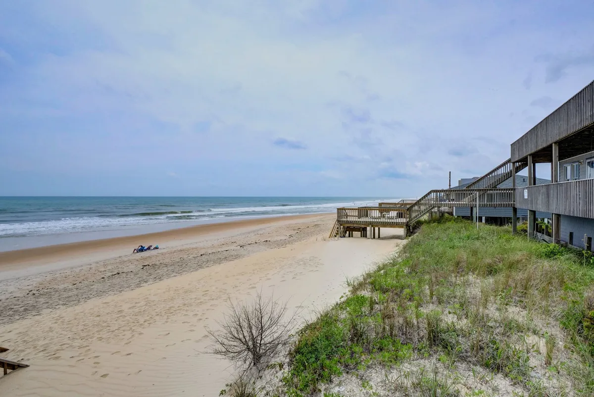 An inviting view of the sandy beach, shimmering sea, and boardwalk bordered by dune vegetation. The scene captures the coastline stretching under a bright sky, suggesting relaxation and outdoor enjoyment.