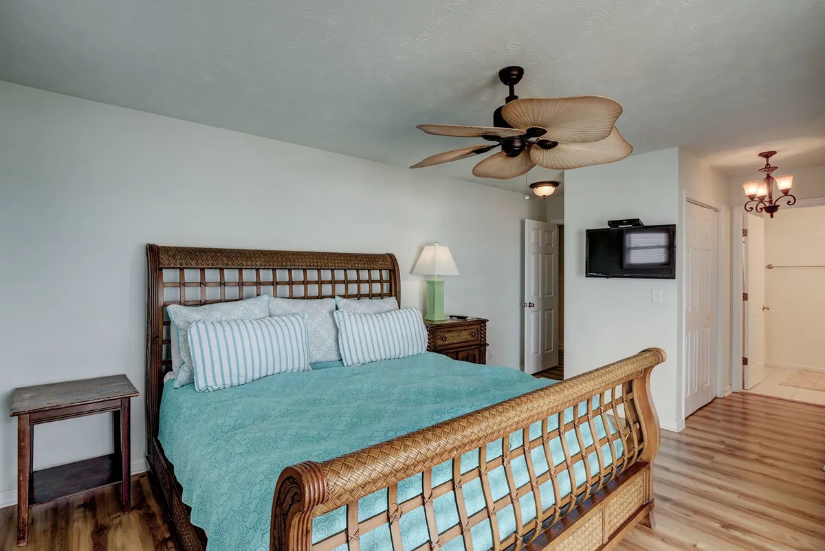 A serene bedroom with a woven headboard bed, striped pillows, a tropical ceiling fan, and a nightstand with a glowing lamp. The airy atmosphere and inviting bedding evoke a sense of peaceful retreat, perfect for shared moments or relaxing mornings. The arrangement suggests easy access to outdoor spaces, aligning with the narrative of direct deck access from the master bedroom.