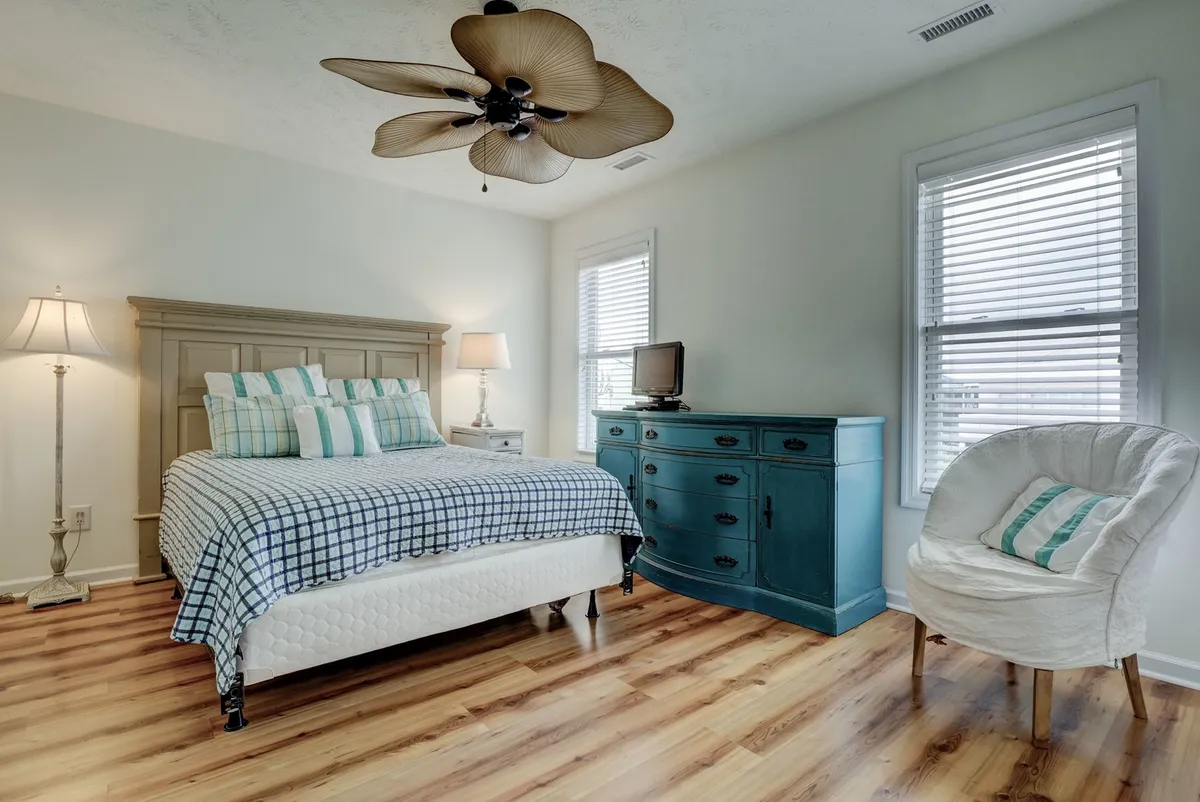 An inviting King bedroom featuring a bed with plaid bedding, a turquoise dresser, a floor lamp for warm lighting, and a comfortable white chair. The space feels intimate and restful, suggesting a quiet place for relaxation or reading.
