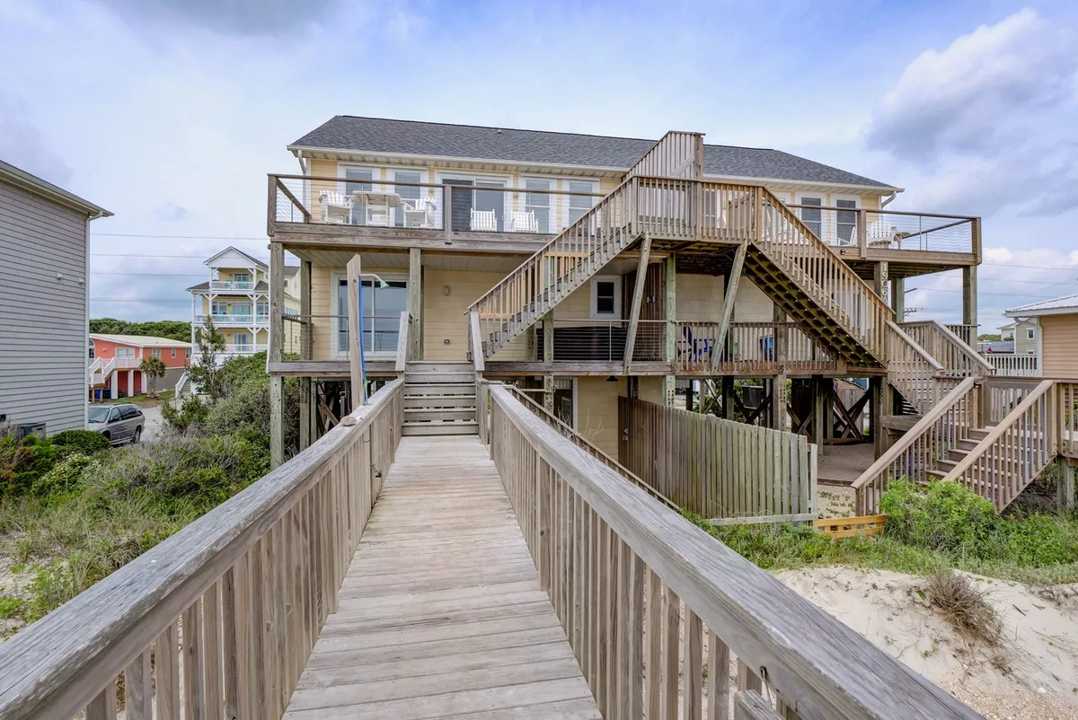 This exterior image shows a wooden walkway leading up to the house, highlighting the multi-level deck surrounded by railings. The house itself is built on stilts, giving it a classic coastal appearance. The outdoor deck is visibly spacious, ideal for social gatherings, relaxing, or letting a pet roam in a secure environment.