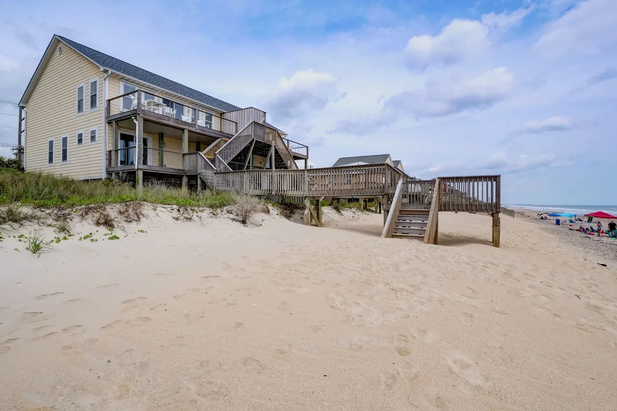 This image shows a beachfront house positioned directly on the sand, with a wooden walkway leading out to the expansive beach. There are beach umbrellas set up, the ocean visible beyond, and a sense of vibrant family activity and relaxation along the shore.