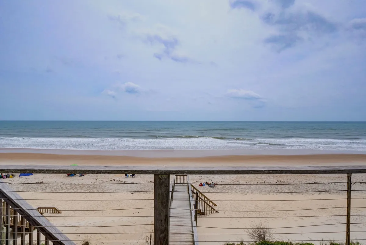 This view captures the broad sandy beach and rolling ocean waves under a cloudy sky. The boardwalk extends along the shore, hinting at local walking opportunities. The atmosphere is serene, inviting morning or evening strolls.