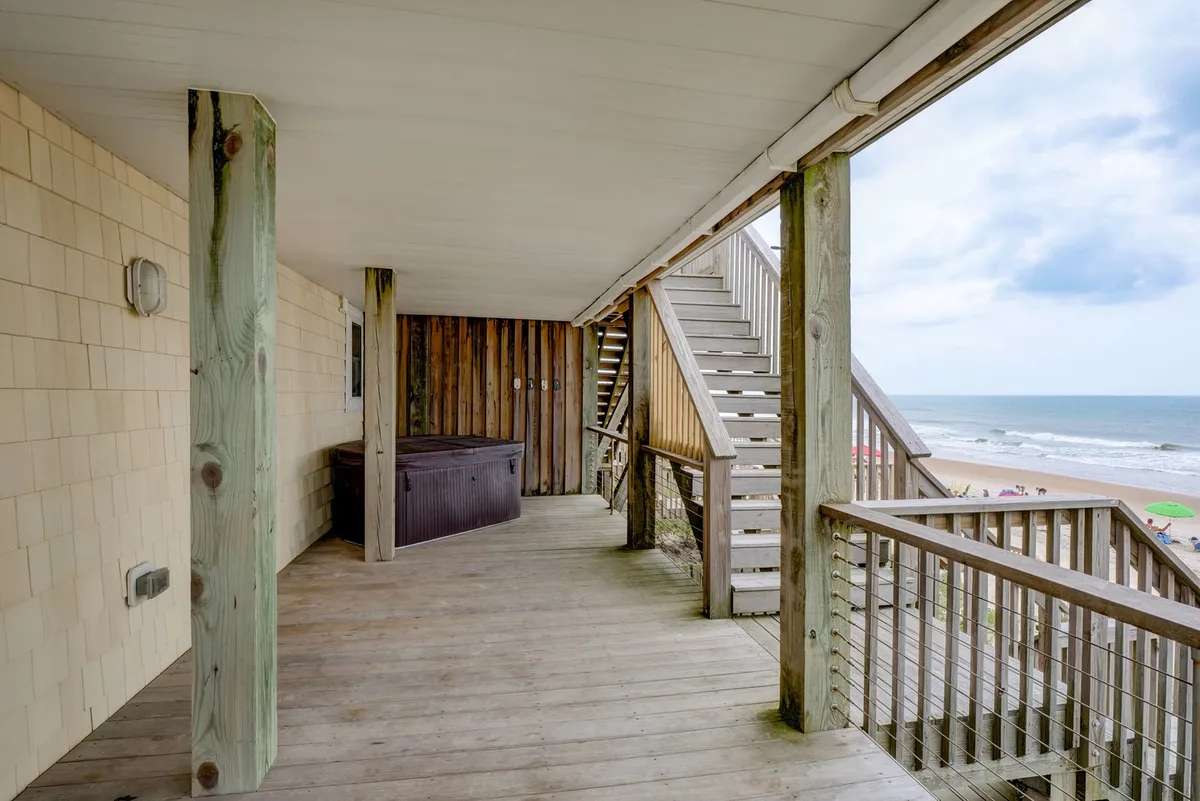 This exterior image features a hot tub situated on a private deck, with a staircase leading down to the beach and a magnificent view of the ocean stretching into the horizon. The modern railing frames the scene, emphasizing the connection between the relaxation of the hot tub and the serene oceanfront backdrop.