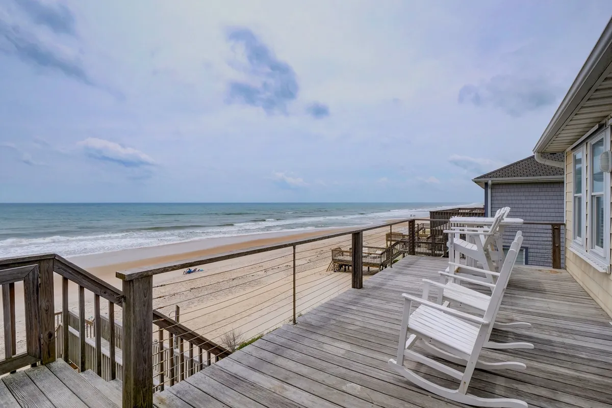 The image showcases a spacious wooden deck overlooking the beach, with inviting lounge chairs perfectly positioned for relaxing and soaking in sweeping ocean views. The scene evokes a sense of tranquility, suggesting sunbathing, reading, or simply enjoying the sound of the waves. Soft morning light or golden hour glow suggests the deck is ideal for both sunrise watching and evening relaxation.