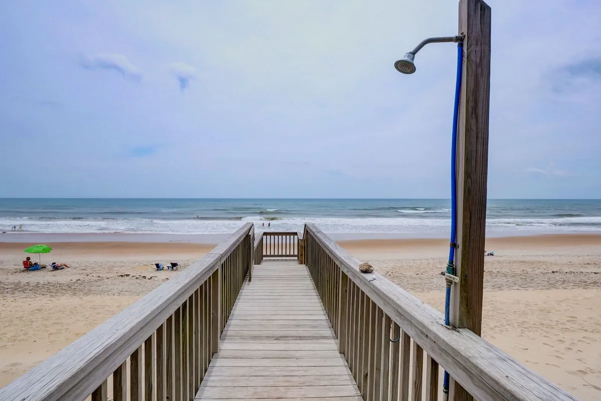An exterior shot featuring a wooden boardwalk that leads directly to the beach, complete with an outdoor shower fixture. The setting is tranquil, conveying a natural transition between home and beach activities.